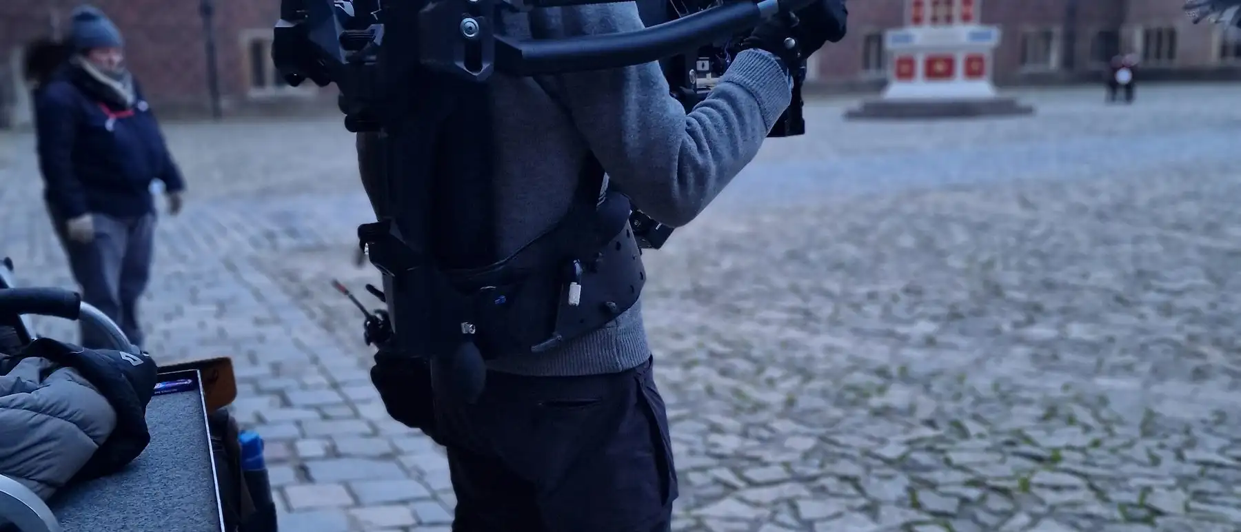An operator carrying a built camera across cobbles on a period location.